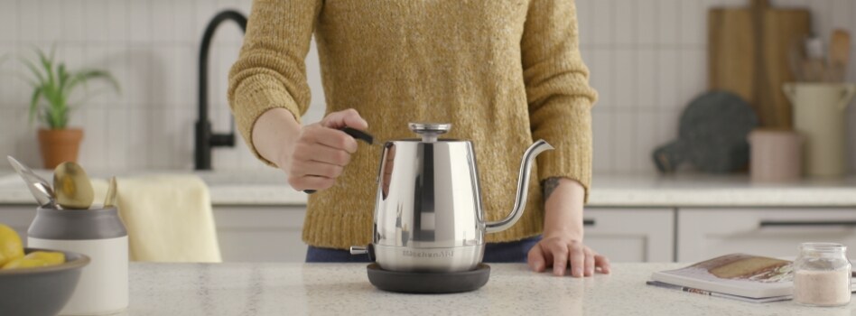 Someone holds the handle of a KitchenAid Gooseneck Kettle that's on a counter. Also on the counter is an opened book and a container of kitchen utensils. In the background is a counter with a sink and various cutting boards and containers.